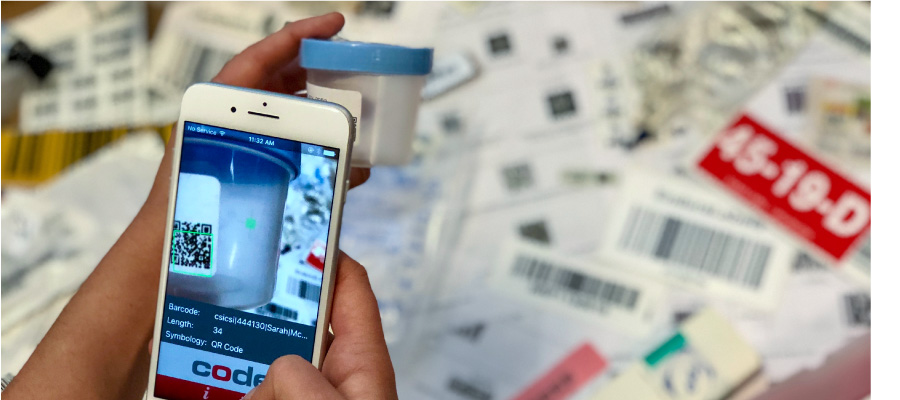 Hand holding phone to scan a sample jar. Multiple barcoded labels in the background.